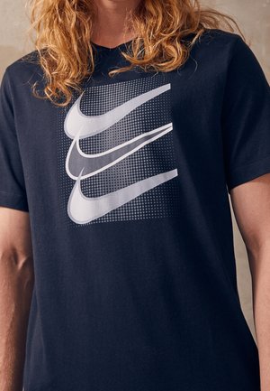 Persoon met lang krullend haar, gekleed in een zwart T-shirt met drie grote overlappende witte swoosh-graphics op een gestippelde achtergrond.