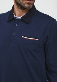 Námornícky modré polo tričko s dlhým rukávom a zapínaním na golieri, s náprsné vreckom a pruhovanými detailami v červenej, bielej a námorníckej modrej farbe.