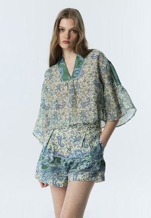 Junge Frau trägt ein lockeres Top mit Paisley- und Blumenmuster, weiten Ärmeln und passenden Shorts, steht mit einer Hand in der Tasche.