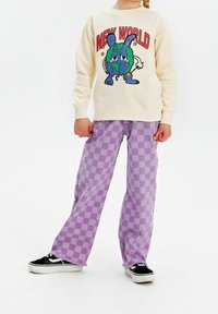 Jasno beżowy sweter z napisem "NOWY ŚWIAT" i grafiką ziemi w stylu kreskówkowym. Fioletowe spodnie w kratę o szerokich nogawkach z postrzępionymi brzegami. Czarne sneakersy.