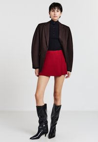 Donna che indossa un blazer marrone scuro, un maglione nero con bottoni, una gonna rossa e stivali neri fino al ginocchio, in piedi davanti a uno sfondo bianco semplice.