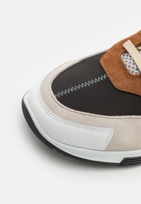 Sneaker z czarną tkaninową cholewką, brązowymi zamszowymi akcentami, beżowymi nakładkami, białą gumową podeszwą oraz kontrastującymi szwami.