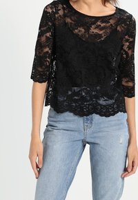 Top in pizzo nero con maniche corte, motivo floreale, orlo ondulato, design a strati. Abbinato a jeans azzurri sdruciti.