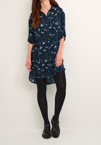 Marineblaues Hemdkleid aus leichtem Stoff mit einem floralen Muster in hellen Farben und einem geschwungenen Saum, kombiniert mit schwarzen Strumpfhosen und Schuhen.
