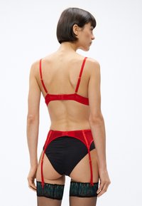 Červená saténová podprsenka s nastaviteľnými ramienkami, v páre s čiernymi nohavičkami s vysokým pásom. Obsahuje podväzkové popruhy a pančuchy s čipkovými akcentmi.