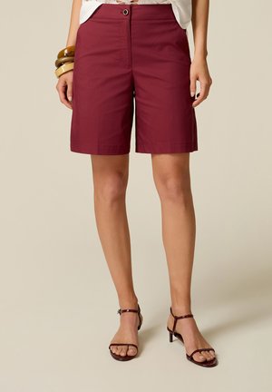 Donna che indossa pantaloncini bordeaux al ginocchio, top bianco, sandali marroni con tacchi alti e tre grandi bracciali al polso sinistro.