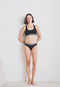Set di reggiseno nero e bikini coordinati con una fascia con logo. Il materiale appare liscio con un design aderente. Il modello è in piedi contro uno sfondo chiaro.