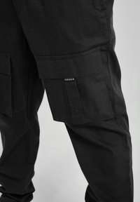 Svarta cargobyxor med stora fickor med klaff och en liten SikSilk-logotyp på höger lårficka mot en enfärgad bakgrund.