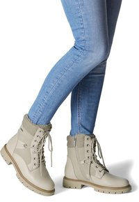Bottes en cuir beige clair avec bordure en fourrure grise, design à lacets et semelles robustes. Portées avec un jean skinny bleu, mettant en valeur la hauteur au niveau de la cheville.