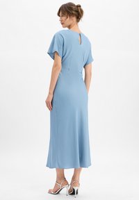 Femme aux cheveux châtain clair relevés en chignon bas, portant une robe midi bleu clair et des sandales à talons hauts argentées, se tenant devant un fond blanc.
