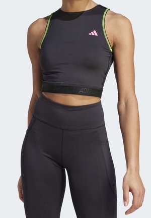 Mujer con un top corto atlético negro con detalles en verde neón y leggings negros de cintura alta, de pie con una postura relajada sobre un fondo claro.