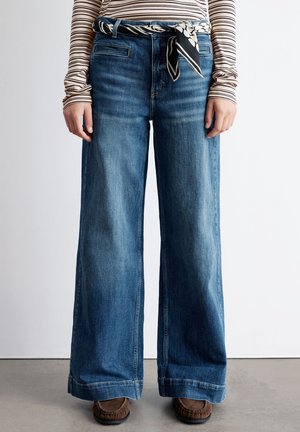 Wide-leg denim jeans in medium blue wash, featuring a high waist, front pockets, and paired with a patterned black and white belt.