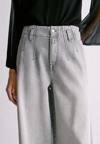 Zbliżenie na osobę w jasnoszarych dżinsach z wysokim stanem, zapinanych na guziki z przodu i z szlufkami na pasek, które są połączone z przezroczystą, czarną bluzką z długim rękawem.