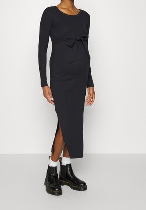 Osoba w ciąży ubrana w dopasowaną czarną sukienkę midi z długim rękawem, z przodu z kokardą i z bocznym rozcięciem, zestawioną z białymi skarpetkami i czarnymi botkami za kostkę.