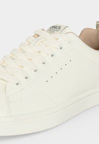 Baskets basses en cuir blanc avec lacets blancs, petits trous de ventilation et doublure intérieure beige. Étiquette de la marque visible sur la languette.