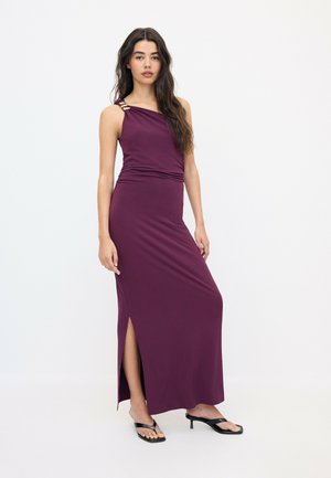 Femme portant une robe sans manches violet foncé avec une fente latérale et des sandales à talons noires, debout devant un fond blanc uni.
