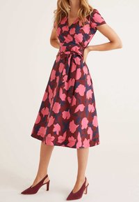 Robe portefeuille en tissu rouge-brun avec un grand motif floral rose et bleu marine ; manches courtes, décolleté en V et taille ceinte. Associée à des talons bordeaux.