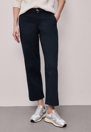 Person iført sorte bukser med lige ben og bælte samt hvide sneakers, den ene hånd i lommen, stående mod en lys væg og gulv.