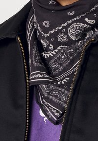 Svart bandana med vita paisleymönster; lättviktigt bomullsmaterial; bärs runt halsen över en lila skjorta, lager med en jacka.