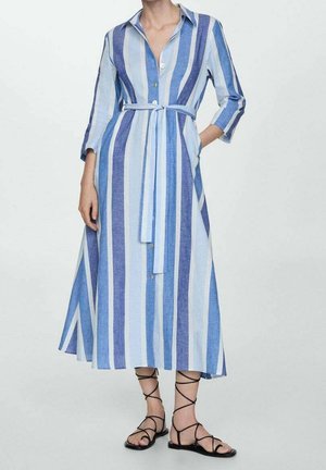Frau trägt ein blau-weiß gestreiftes Midi-Kleid mit Gürtel, Knopfleiste vorne, Dreiviertelärmeln und schwarzen flachen Sandalen mit Schnürung.