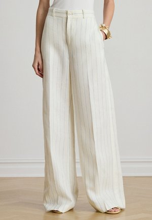 Naine kannab kõrge vöökohaga, laia säärega kreemikaid pükse õhukeste vertikaalsete siniste triipudega, mille juurde kuuluvad beežid avatud varbaga sandaalid ja kuldne käevõru.