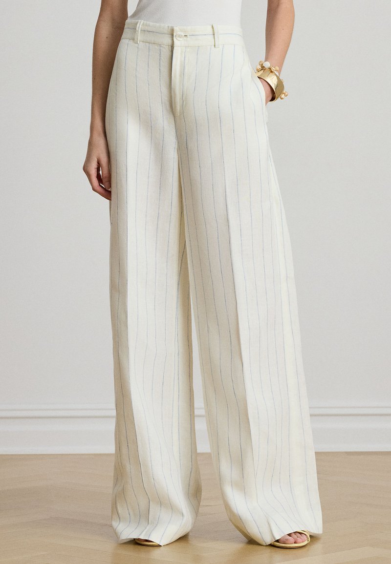 Nő, aki magas derekú, széles szárú krémszínű nadrágot visel vékony, függőleges kék csíkokkal, bézs nyitott orrú szandállal és arany karkötővel.