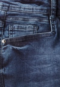 Tissu en denim bleu foncé avec une texture légèrement décolorée, comportant une poche arrière, un rivet argenté et des détails de couture contrastants.