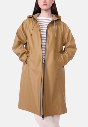 Donna che indossa un impermeabile color beige con cappuccio, cerniera frontale, coulisse e tasche, sopra una camicia a righe bianche, in piedi con le mani in tasca.