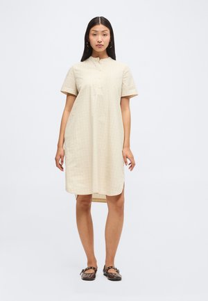 Femme debout portant une robe beige mi-longue à manches courtes avec des boutons sur le devant et des chaussures Mary Jane foncées.