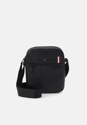 Kompakt sort crossbody-taske med justerbar rem, frontlomme med lynlås og Tommy Hilfiger-logo med rød, hvid og blå stribe.