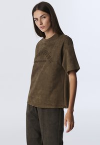 Bruin suède shirt met korte mouwen en een ronde halslijn. Geweven oppervlak en reliëflettering op de voorkant. Eenvoudige, losse silhouet.