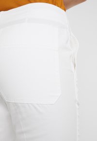 Bílé bavlněné kalhoty mají hladkou, jemnou texturu, boční kapsu a čistý design pasu. Materiál se jeví jako lehký a odolný.