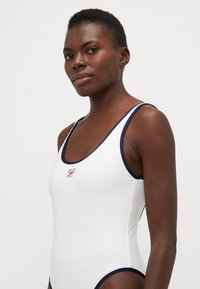Witte eendelige zwemkleding met marineblauwe accenten, voorzien van een ronde halslijn en dunne bandjes. Speedo-logo in het rood nabij de borst.