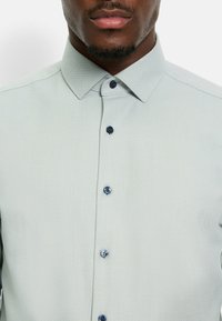 Helles grünes, gemustertes Hemd mit geometrischem Design, spitzem Kragen und dunkelblauen Knöpfen entlang der Vorderseite. Strukturiertes Material.