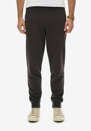 Mand iført mørkegrå joggerbukser med elastiske manchetter og beige sneakers, stående mod en hvid baggrund.