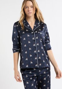 Granatowy satynowy zestaw piżamowy z białym pipingiem, zdobiony zabawnym wzorem z psami. Koszula ma kołnierzyk i kieszeń; spodnie mają prostą nogawkę.