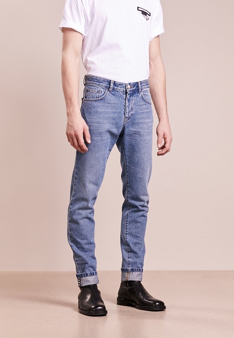 Man i ljusblå uppvikta jeans, vit t-shirt med kort ärm och svarta läderstövlar som står på en beige golv.