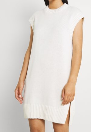 Sleeveless white knit dress with ribbed crew neck, cuffs, and hem, featuring side slits, worn by a standing person.