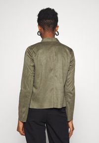 Giacca in suede verde oliva con una vestibilità su misura, maniche lunghe e colletto strutturato. Texture liscia, linee pulite e dettaglio della cucitura posteriore.