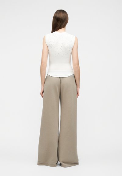 Μια γυναίκα στέκεται γυρισμένη με την πλάτη, φοράει αμάνικο λευκό τοπ με μικρά τρουκ και φαρδιά μπεζ παντελόνια.