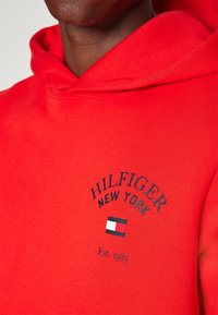 Rode hoodie gemaakt van zachte stof, met geborduurd logo "HILFIGER NEW YORK" boven een klein vlagembleem. Inclusief detail "Opgericht in 1985".