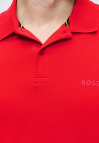 Gros plan d'un polo rouge avec col, un bouton fermé, et un logo discret "BOSS" sur la poitrine.