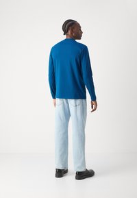 Langermet blå skjorte med rund hals, sett fra baksiden, kombinert med lyseblå jeans og svart fottøy. Jevn stofftekstur.