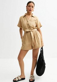 Combinaison en lin beige à manches courtes avec un col, fermeture à boutons sur le devant, taille ceintrée et poches latérales. Sac à main tissé noir et sandales avec boucles.
