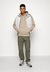Light gray padded vest over beige hoodie with drawstrings, olive green cargo pants, and black and white sneakers with red accents.
