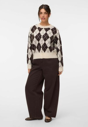 Kvinde iført en hvid og mørkebrun argyle cardigan, mørke vidde bukser og mønstrede flade sko, stående mod en ensfarvet baggrund.