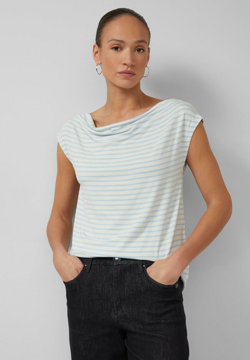 Vrouw met haar naar achteren gebonden, draagt een lichtblauw-wit gestreepte mouwloze top en zwarte broek, handen in de zakken, neutrale gezichtsuitdrukking.