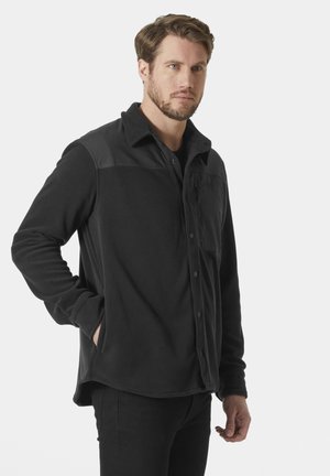 Hombre con cabello castaño claro y barba, vistiendo una chaqueta de forro polar negra con cuello, de pie con una mano en el bolsillo contra un fondo blanco.