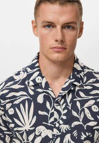 Camisa de manga corta hecha de una tela ligera, con un fondo azul marino y patrones de hojas tropicales y flores en blanco, y un cuello tipo polo.
