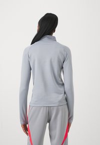 Top sportivo grigio a maniche lunghe con collo alto, texture liscia e design aderente. Presenta dettagli rosa sulle maniche e nei pannelli laterali.